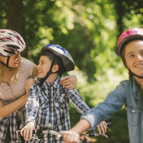Family riding bicycle in the public park together. Parents putting cycling helmet on childrens head
